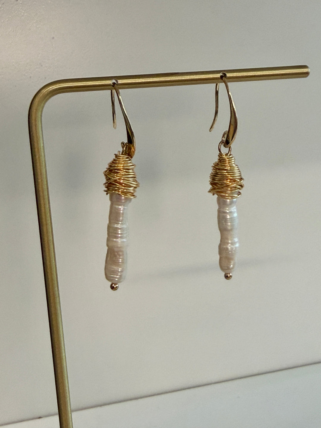 A pair of handmade dangle earrings hanging from a gold metal display stand against a plain light background. Each earring has a gold fishhook back, a wrapped gold top section, and a vertical baroque pearl bead dangling below.