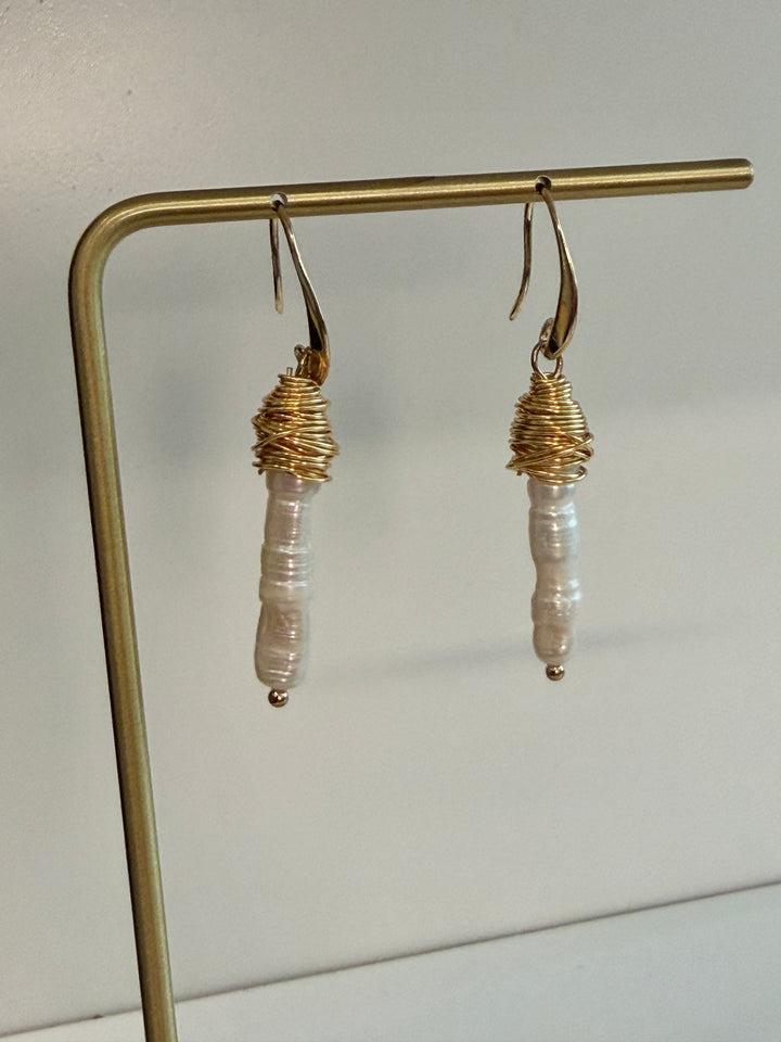 A pair of handmade dangle earrings hanging from a gold metal display stand against a plain light background. Each earring has a gold fishhook back, a wrapped gold top section, and a vertical baroque pearl bead dangling below.